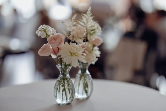 Zwei Glasvasen mit zartrosa und weißen Blumen auf einem Tisch.